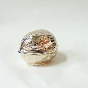 May include: A metallic, gold-toned trinket box shaped like a walnut. The object features a highly reflective, polished surface with intricate, embossed textures that mimic the natural, irregular ridges and grooves found on a real walnut shell.