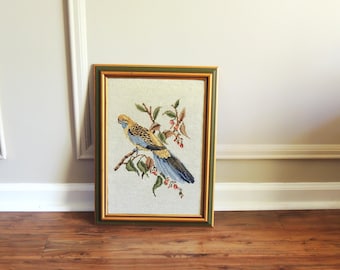 May include: Framed needlepoint art featuring a colorful bird perched on a branch with leaves and berries. The bird has yellow, blue, and black feathers. The frame is gold and green, adding a vintage touch to the artwork.