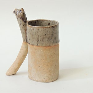 K&ouml;nnte beinhalten: Eine Keramiktasse mit einem einzigartigen Griff in Form eines Tierkopfes. Die Tasse ist in einem hellbraunen Ton glasiert, mit einem dunkleren braunen Band um den oberen Rand.