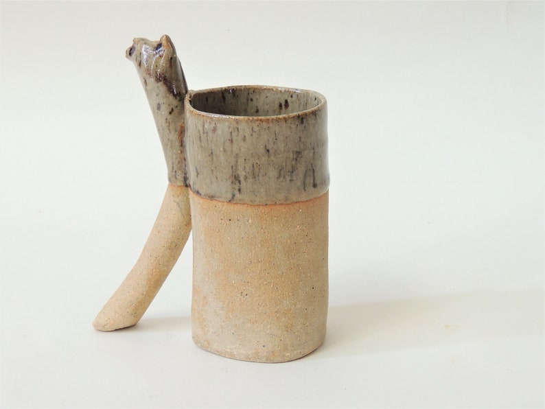 K&ouml;nnte beinhalten: Eine Keramiktasse mit einem einzigartigen Griff in Form eines Tierkopfes. Die Tasse ist in einem hellbraunen Ton glasiert, mit einem dunkleren braunen Band um den oberen Rand.