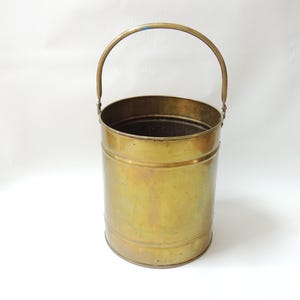 May include: A vintage brass bucket with a rounded handle. The bucket has a cylindrical shape with horizontal bands around the body. The brass has a warm, aged patina, suggesting it's an antique or a well-loved item. The handle is attached to the top rim.