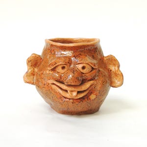 Könnte beinhalten: Eine braune Keramiktasse in Form eines Gesichts mit einem breiten Lächeln und einer herausgestreckten Zunge. Die Tasse hat braune Flecken in der gesamten Glasur.