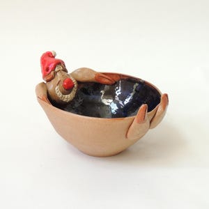 May include: A whimsical ceramic bowl with a figure of a person wearing a red hat and a red nose. The bowl is tan with a dark blue interior. The figure is resting in the bowl, with its arms and legs extending over the sides.