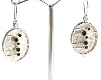 Puede incluir: Un par de pendientes de plata con colgantes ovalados de acabado cepillado y texturizado. Cada colgante presenta una serie de pequeñas hendiduras circulares dispuestas en línea vertical, suspendidos de sencillos ganchos de plata para las orejas.