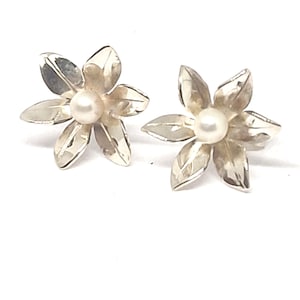May include: A pair of stud earrings shaped like six-petaled flowers. Each earring is crafted from a polished silver-toned metal and features a small, round, white pearl set in the center of the flower petals, creating a floral design.