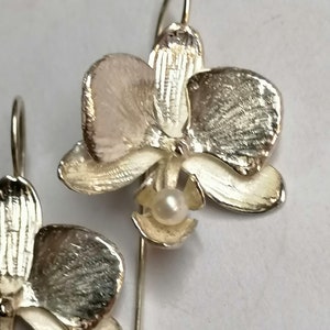 May include: A pair of silver orchid earrings with a pearl accent. The earrings are detailed with textured petals and a delicate stem.