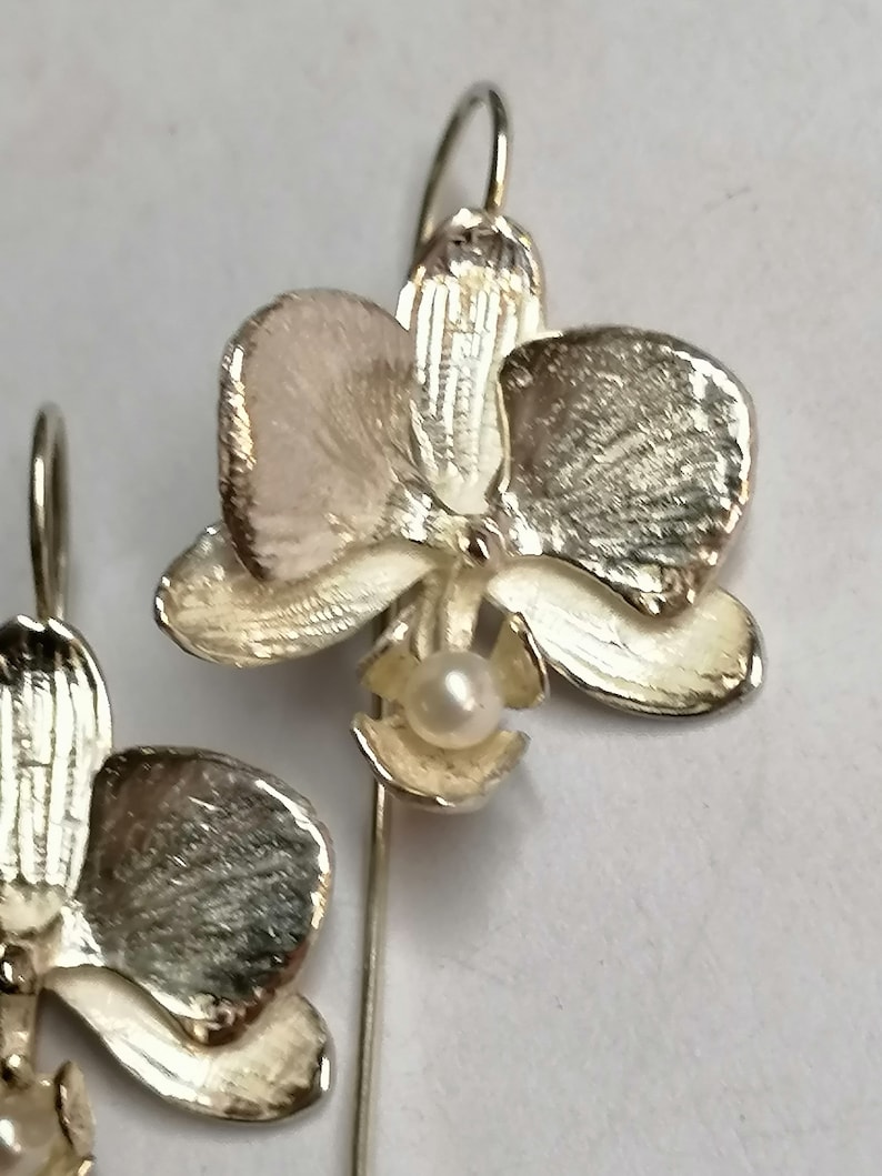 May include: A pair of silver orchid earrings with a pearl accent. The earrings are detailed with textured petals and a delicate stem.