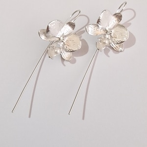 May include: A pair of silver orchid earrings with a pearl accent. The earrings are long and dangle.