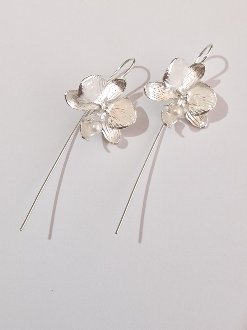 May include: A pair of silver orchid earrings with a pearl accent. The earrings are long and dangle.