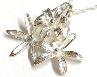 Puede incluir: Un collar con colgante de plata que presenta tres flores tridimensionales con pétalos texturizados y un estambre esférico central en cada flor. El colgante está unido a una delicada cadena de plata, con un acabado metálico pulido.