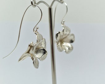 May include: A pair of silver dangle earrings featuring a delicate flower design. The earrings are hanging from a silver stand.