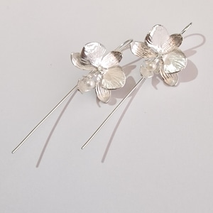 May include: A pair of silver dangle earrings featuring a delicate orchid flower design with a small pearl accent. The earrings have a long, thin wire that hangs down from the flower.