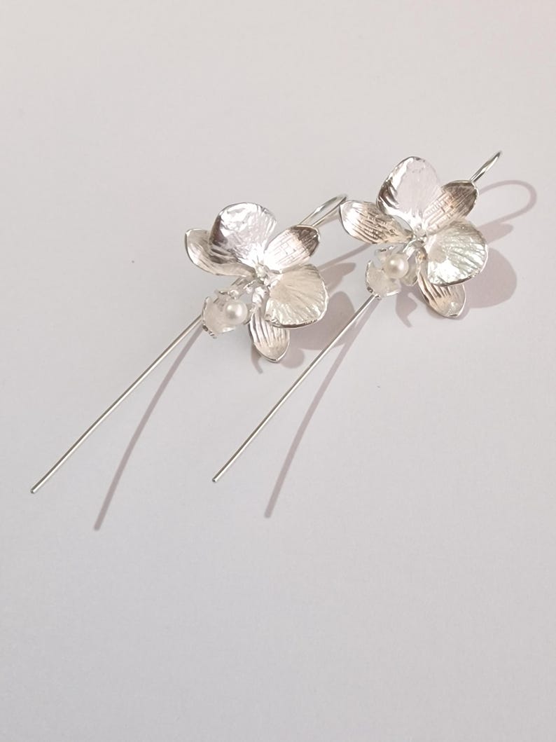 May include: A pair of silver dangle earrings featuring a delicate orchid flower design with a small pearl accent. The earrings have a long, thin wire that hangs down from the flower.