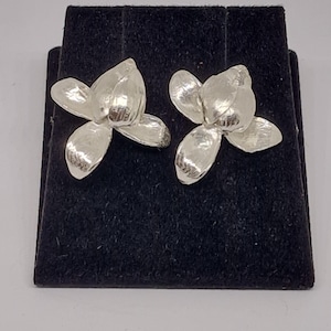 Op de afbeelding: Een paar zilverkleurige bloemvormige oorstekers. Elke oorbel heeft drie bloemblaadjes, waardoor een bloemmotief ontstaat. De oorbellen worden gepresenteerd op een zwart fluwelen oppervlak, waardoor hun metallic glans wordt benadrukt. Het zijn sieraden.