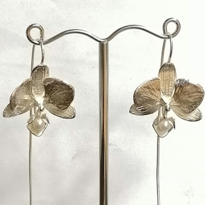 May include: A pair of silver orchid earrings with a pearl accent hanging from a silver hook.