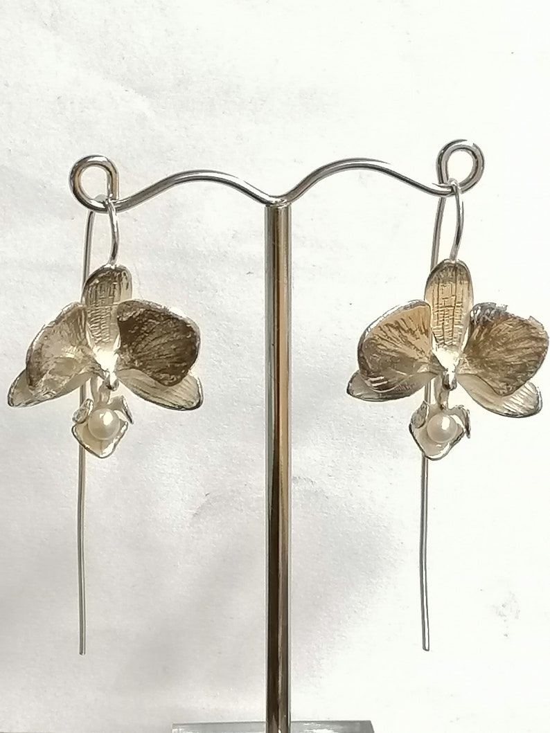 May include: A pair of silver orchid earrings with a pearl accent hanging from a silver hook.
