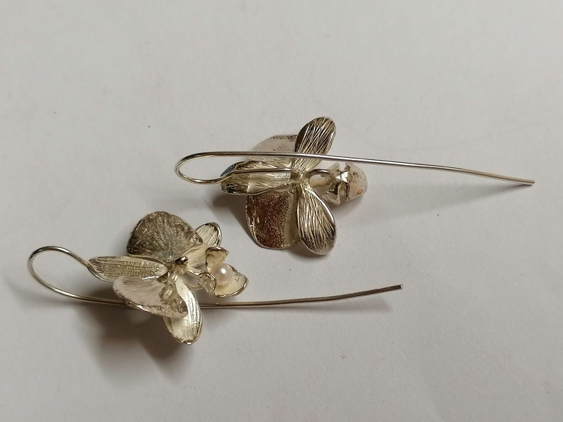 May include: A pair of silver orchid earrings with a pearl accent. The earrings are dangle style with a delicate wire.