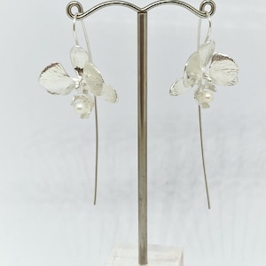 May include: A pair of silver orchid earrings with a pearl accent hanging from a silver stand.