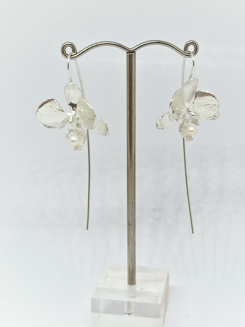 May include: A pair of silver orchid earrings with a pearl accent hanging from a silver stand.