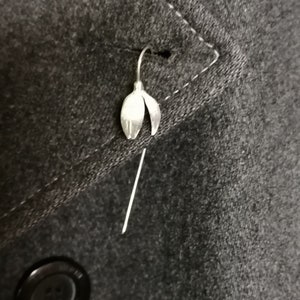 May include: A silver snowdrop flower brooch pin on a dark gray fabric background.