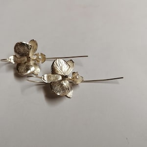 May include: A pair of silver orchid earrings with a pearl accent. The earrings are dangle style with a delicate wire.