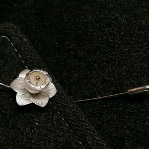 Puede incluir: Un broche de flor de plata con un centro dorado. El broche est&aacute; unido a un alfiler de plata.