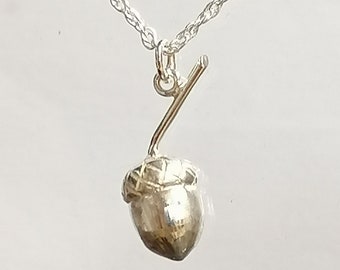 Puede incluir: Un collar con un colgante de bellota de plata en una delicada cadena de cuerda. La bellota está detallada con una superficie texturizada y un pequeño tallo.