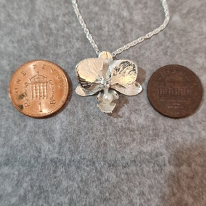 May include: A silver orchid pendant necklace with a pearl accent. The necklace is shown next to two British coins for size comparison.
