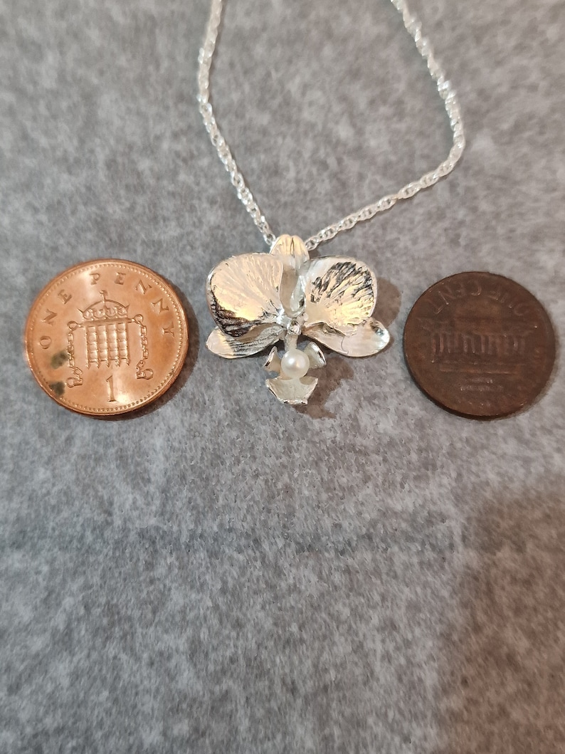 May include: A silver orchid pendant necklace with a pearl accent. The necklace is shown next to two British coins for size comparison.