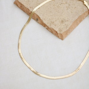 May include: A gold-colored herringbone chain necklace. The necklace is displayed on a textured, light brown stone against a white background. The chain has a flat, woven design and a clasp closure.