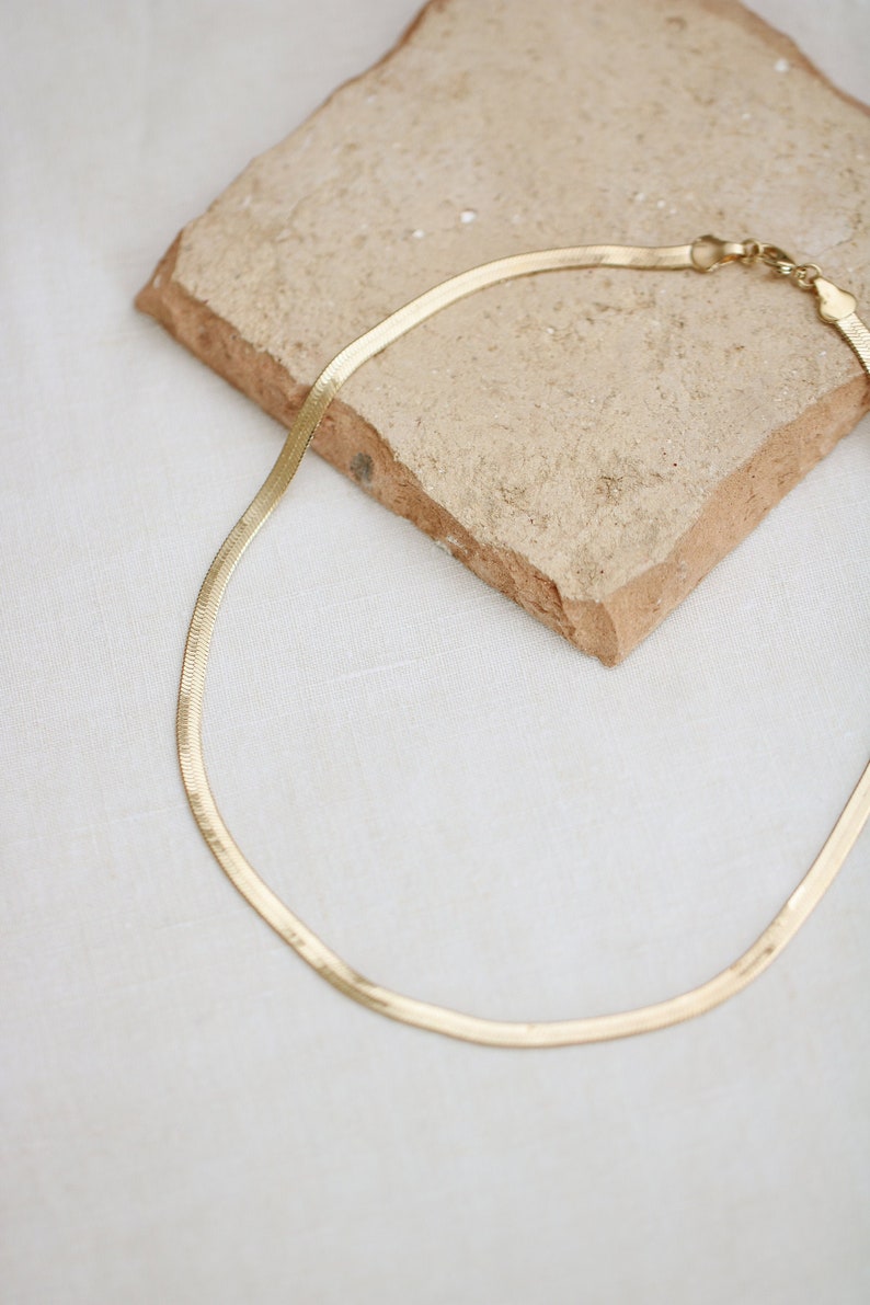 May include: A gold-colored herringbone chain necklace. The necklace is displayed on a textured, light brown stone against a white background. The chain has a flat, woven design and a clasp closure.