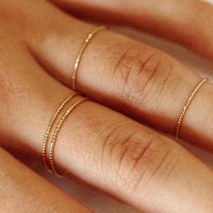 Peut inclure: Gros plan sur plusieurs bagues en or délicates. Deux bagues sont empilées sur un doigt, tandis que d'autres sont portées sur les doigts adjacents. Les bagues ont un design texturé et perlé, et sont de couleur dorée chaude.