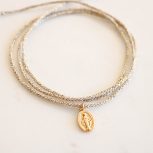 Peut inclure: Un bracelet délicat avec un pendentif en or représentant la Vierge Marie sur un cordon argenté.