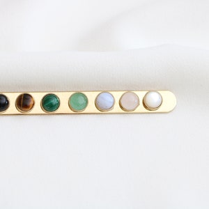 May include: A gold bar with seven round gemstones set in it. The gemstones are black, brown, green, green, light blue, pink, and white.