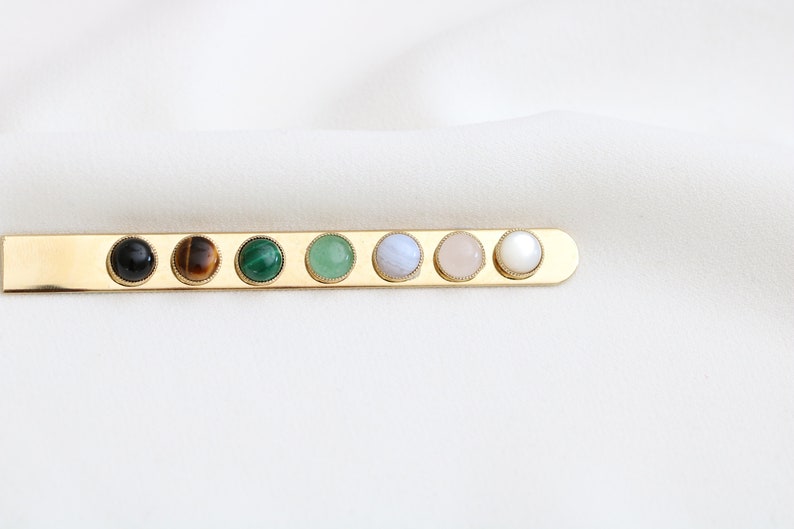 May include: A gold bar with seven round gemstones set in it. The gemstones are black, brown, green, green, light blue, pink, and white.