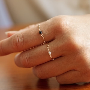 Puede incluir: Dos delicados anillos de cadena de oro con una sola piedra preciosa negra y una sola perla blanca.