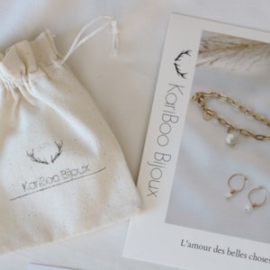 May include: An off-white drawstring bag with the text "KariBoo Bijoux" and a gold-colored bracelet, necklace, and earrings with white pearl accents. A card with the text "L'amour des belles choses" is also visible.