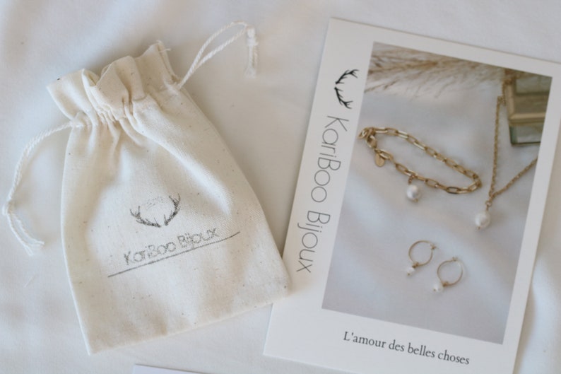 May include: An off-white drawstring bag with the text "KariBoo Bijoux" and a gold-colored bracelet, necklace, and earrings with white pearl accents. A card with the text "L'amour des belles choses" is also visible.