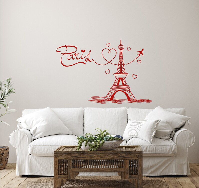 May include: Red Eiffel Tower wall decal with the word "Paris" and hearts. The decal is on a white wall above a white couch with a wooden coffee table.