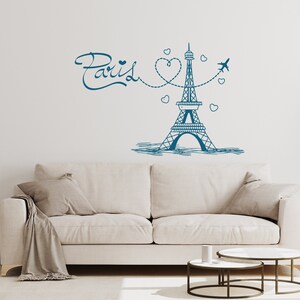 May include: A white couch with two pillows and a throw blanket sits in front of a white wall with a blue and white illustration of the Eiffel Tower, a heart, and the word "Paris".