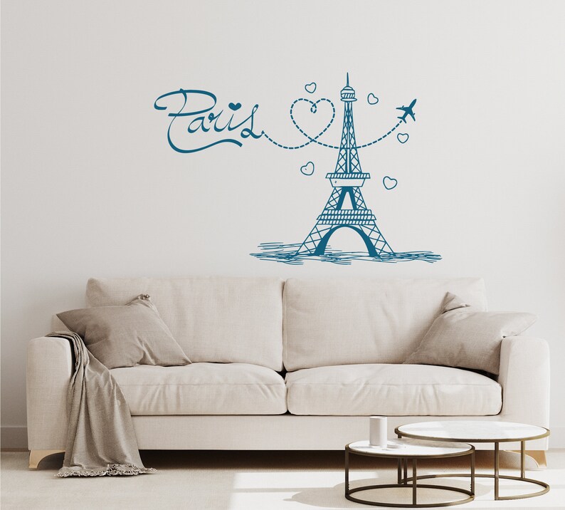 May include: A white couch with two pillows and a throw blanket sits in front of a white wall with a blue and white illustration of the Eiffel Tower, a heart, and the word "Paris".