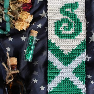May include: A green and white crocheted wall hanging with the letter "S" in the center. The hanging is made with a white background and green yarn.