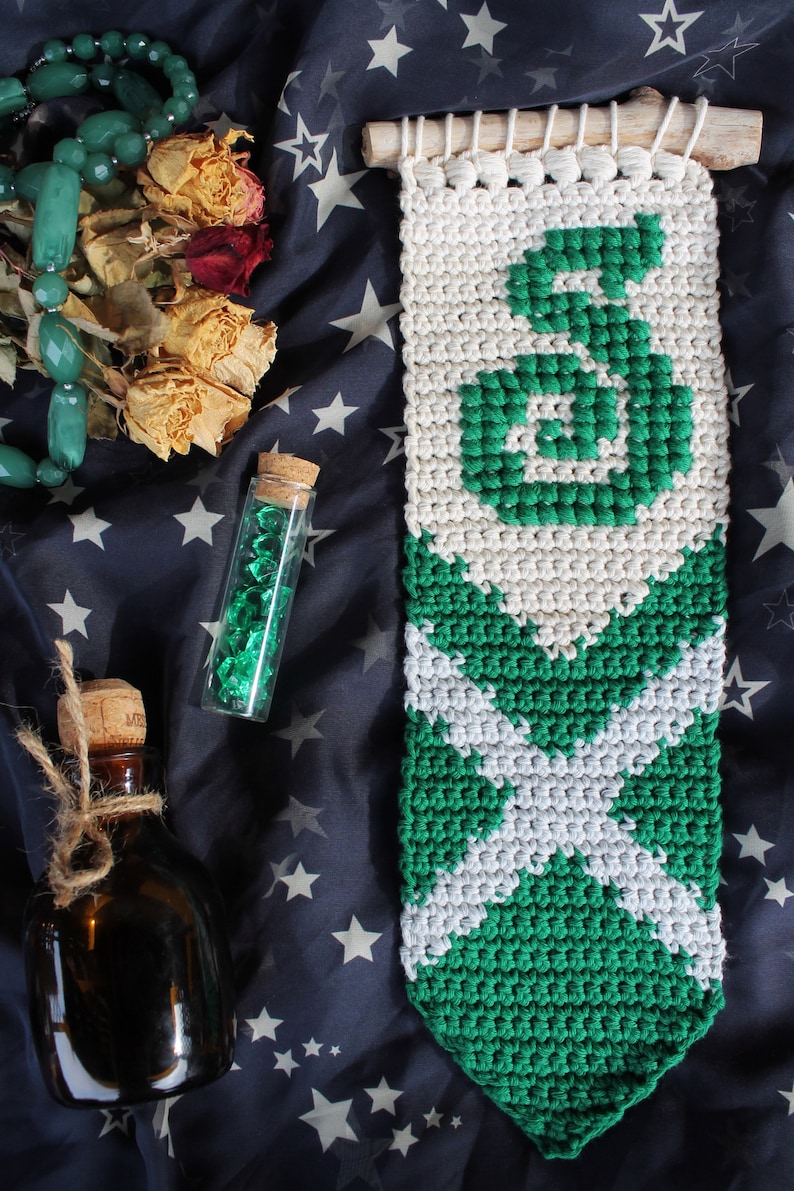 May include: A green and white crocheted wall hanging with the letter "S" in the center. The hanging is made with a white background and green yarn.
