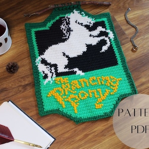 Può includere: Un arazzo da parete all'uncinetto verde, nero e bianco con un disegno di cavallo e le parole "The Prancing Pony" in giallo. Lo stendardo è esposto su un'asta di legno. L'immagine include anche una tazza, una pigna e un taccuino.