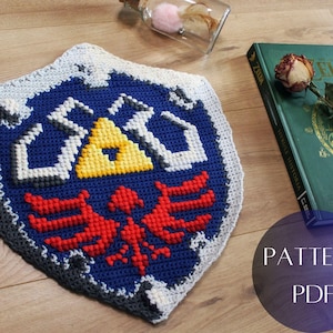 Pode incluir: Um escudo de crochê feito à mão no estilo de The Legend of Zelda. O escudo apresenta um fundo azul com um triângulo amarelo e um emblema de pássaro vermelho. A borda é cinza e branca.