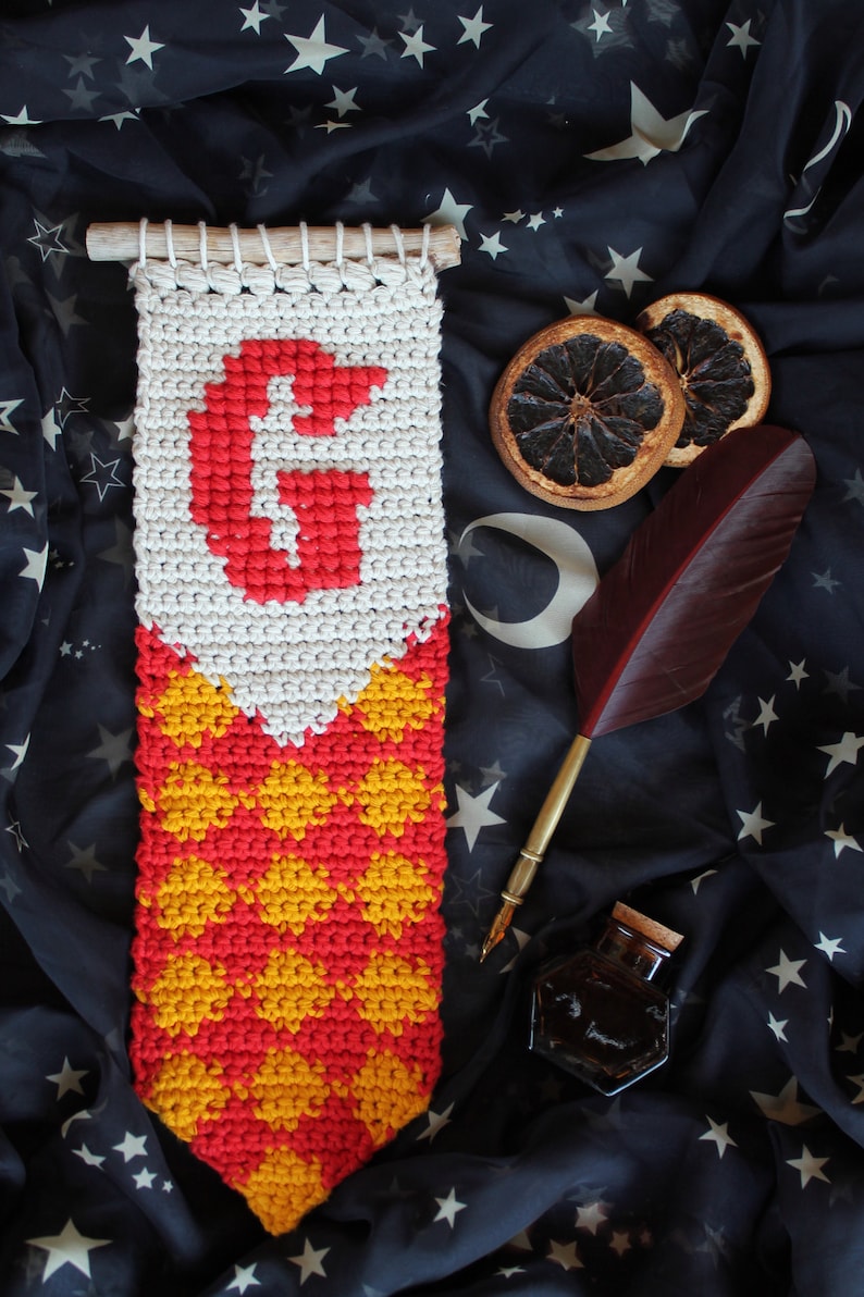 May include: A crocheted tapestry with a red "G" and a red and yellow checkered pattern. The tapestry is hanging on a black fabric background with stars.