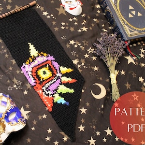 May include: A crochet wall hanging featuring a colorful, pixelated design of a video game character. The hanging is black with a wooden dowel at the top. The text "PATTERN PDF" is visible in the lower right corner.