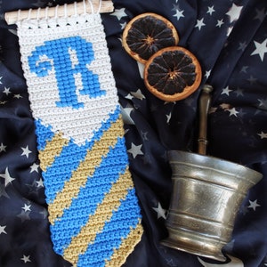 May include: A crocheted wall hanging with a blue and gold striped design and a white section with the letter "R" in blue. The hanging is on a dark blue fabric with white stars.