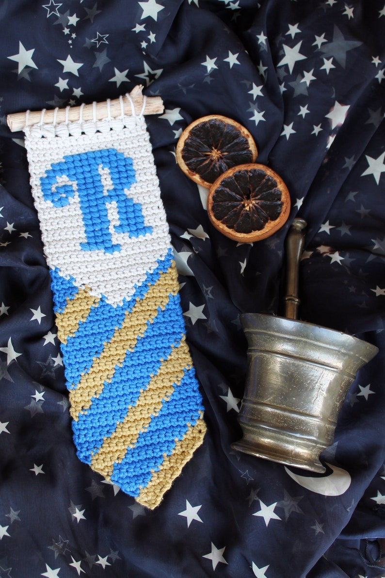 May include: A crocheted wall hanging with a blue and gold striped design and a white section with the letter "R" in blue. The hanging is on a dark blue fabric with white stars.