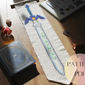 May include: A handmade, off-white crochet bookmark featuring a blue and gold sword design with green text. The bookmark is lying on a wooden surface next to a book and other decorative items. The words "PATTERN PDF" are in a pink circle.